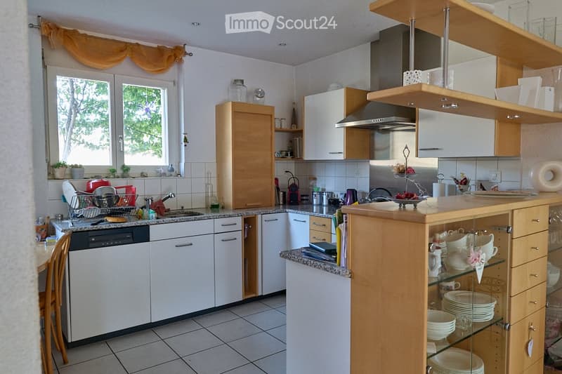 Modern kitchen with white and wooden cabinets, granite countertops, a central island, and various appliances including a dishwasher, microwave, and toaster. Shelves and open storage display dishes and cups.
