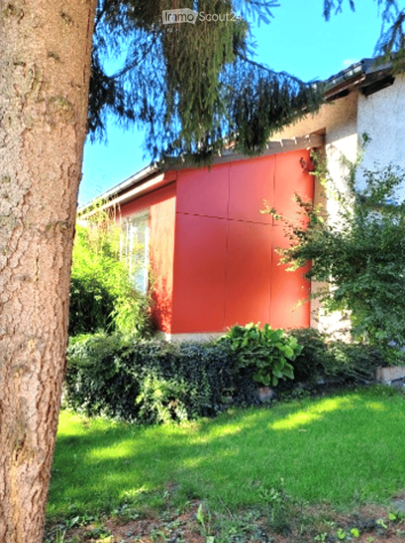 A modern, single-story house with a red facade and white trim, surrounded by a well-maintained garden and trees.
