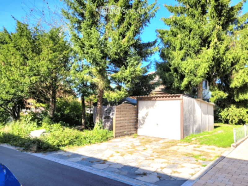A well-maintained garden area with a paved driveway leading to a garage, surrounded by lush green trees and shrubs.