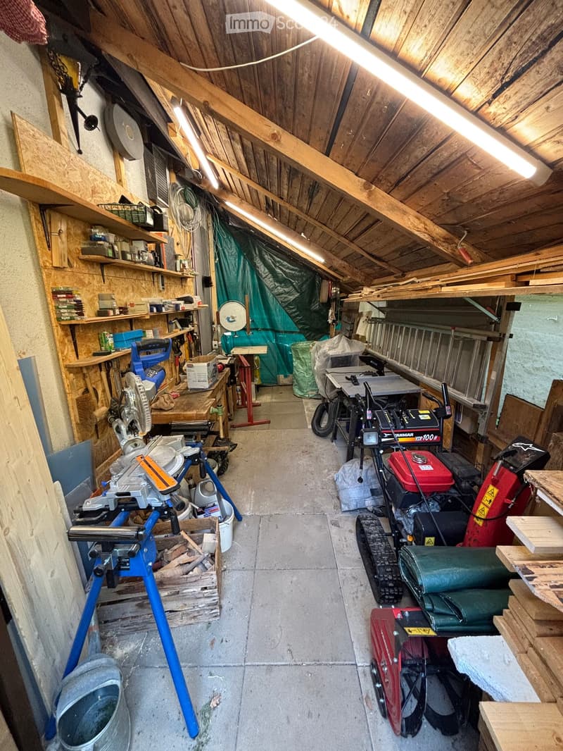 Workshop with exposed wooden beams, concrete floor, and various tools and equipment including a saw, table, shelves, and a lawn mower.