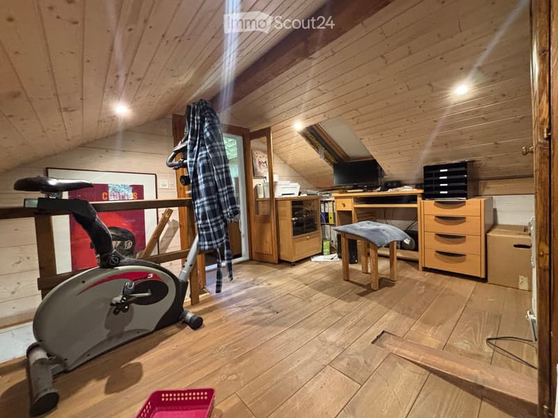 A cozy attic room with wooden walls and ceiling, featuring an exercise bike, a small desk with a chair, a cabinet, and a screen. A plaid blanket is draped over a chair, and a framed picture hangs on the wall.