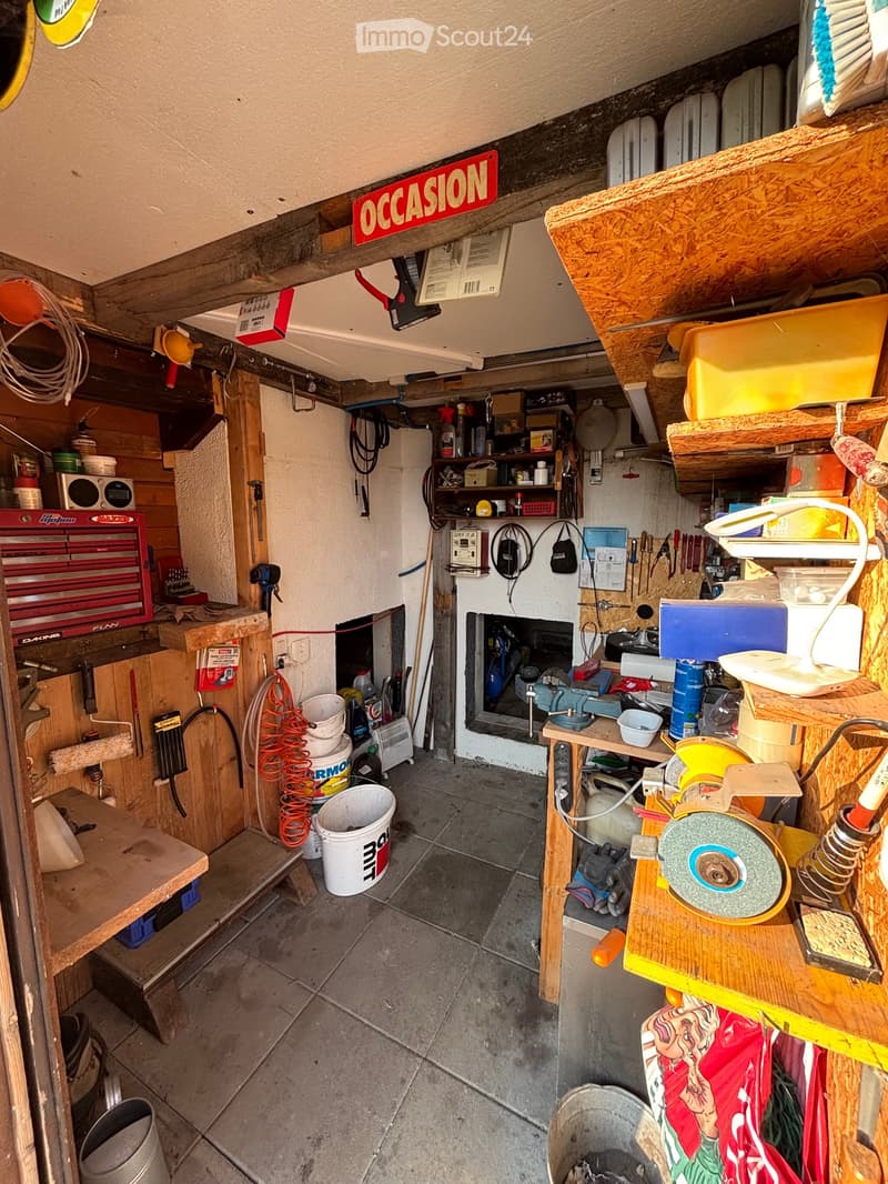 A cluttered garage or workshop space with various tools and equipment, including a red toolbox, a grinder on a workbench, shelves with tools, and a sign that reads "OCCASION".