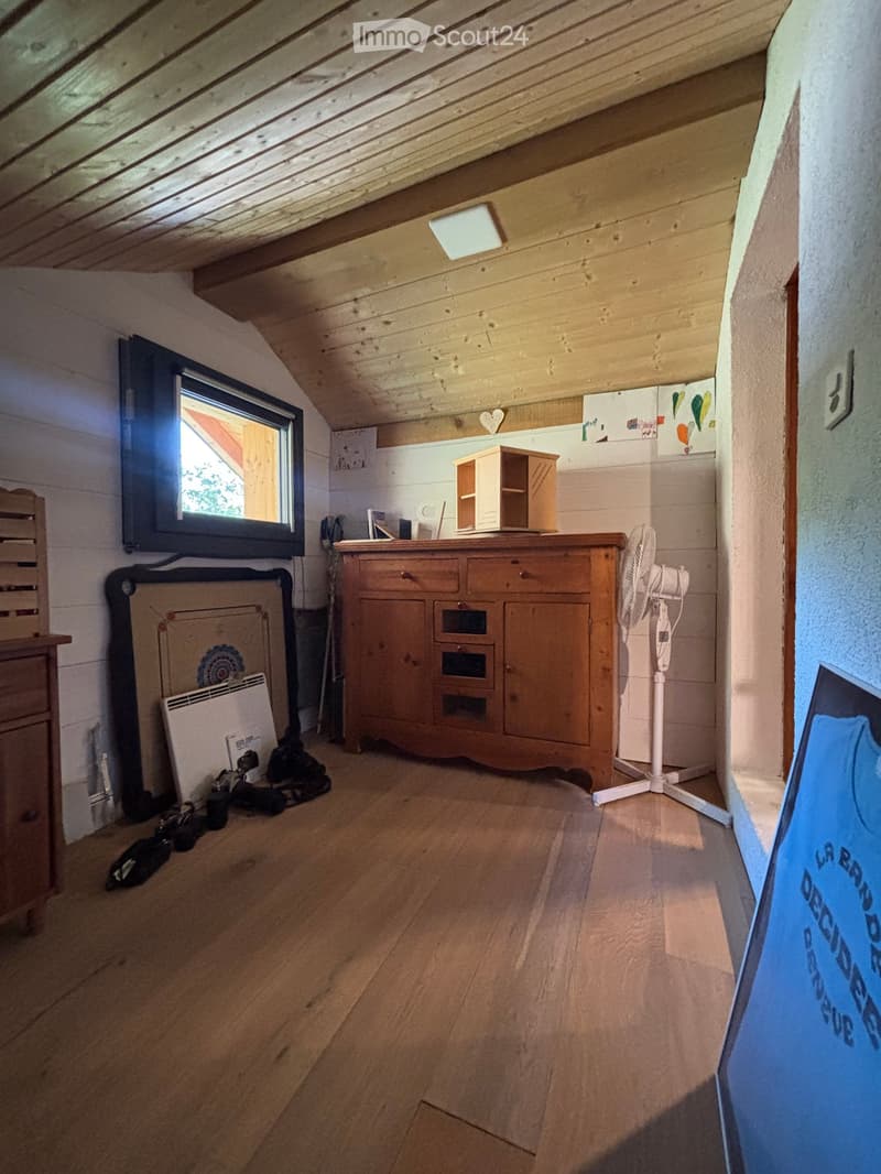 Attic room with wooden slanted ceiling, exposed beams, wooden floor, window, cabinet, fan, mirror, radiator, and shelf.