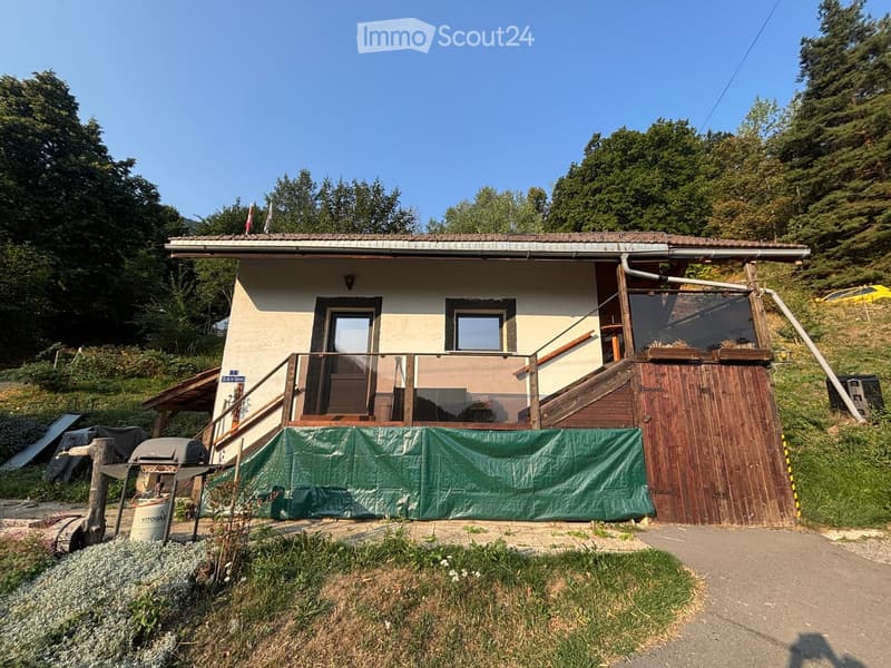 A small, single-story house with a tiled roof, beige walls, and a wooden balcony. The house has a covered entrance and is surrounded by greenery and trees.