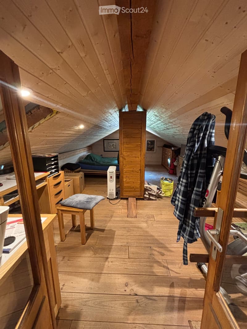 Cozy attic room with wooden walls and slanted ceilings, featuring a bed, wardrobe, small table, and a heater.