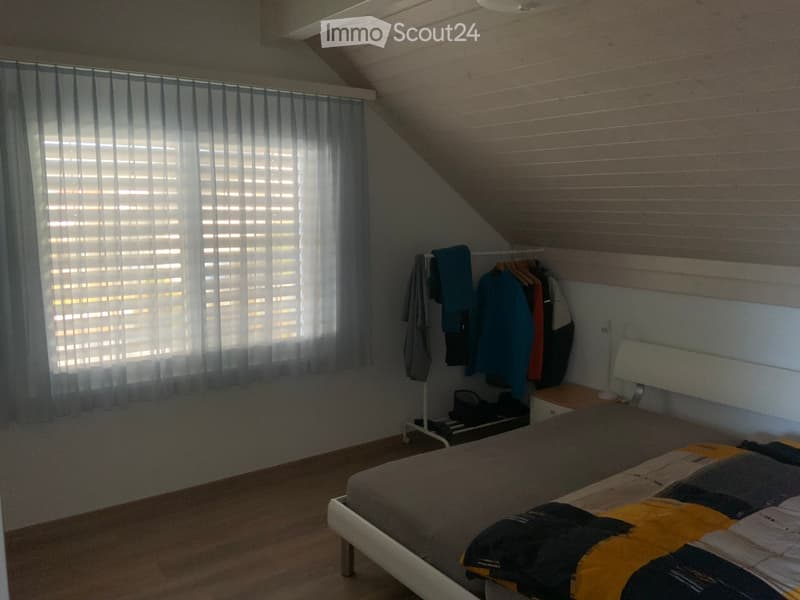 white walls, bed with duvet, wooden floor, window with gray curtains, white headboard, nightstand, clothes rack with folded clothes