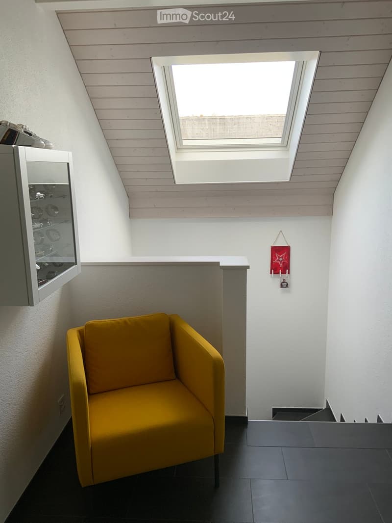 A room with a white wall, black floor, a yellow armchair, and a display case with glass. It also has a skylight and a staircase.