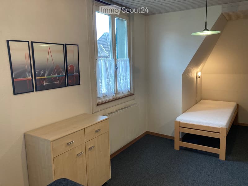A small bedroom with a single bed, a wooden cabinet, a radiator, and three framed mirrors on the wall. The room has a window with white curtains and a pendant light fixture.