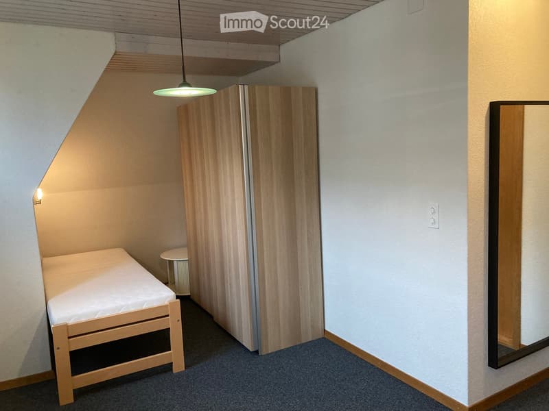 Small bedroom with a single bed, a small stool, and a wooden wardrobe. The room has a slanted ceiling and is well-lit with a hanging light fixture.