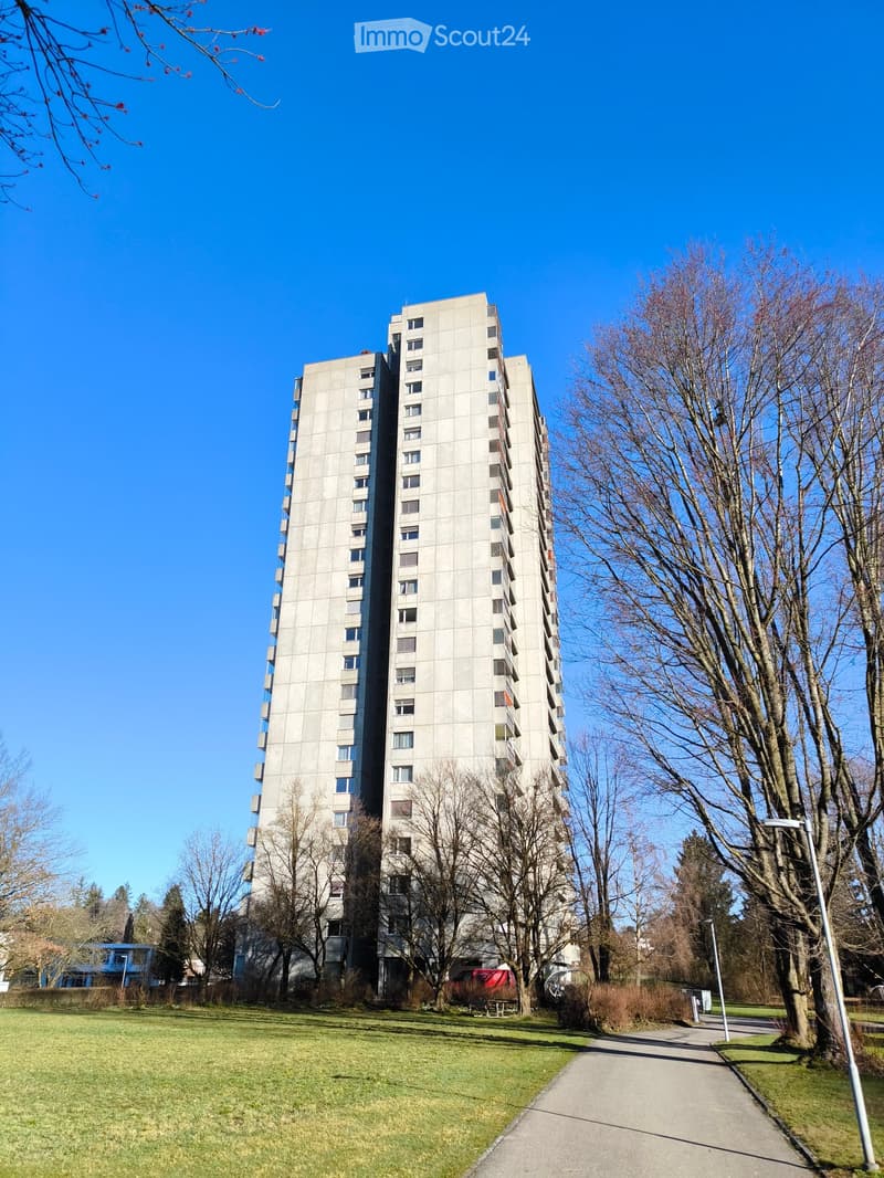 High-rise building with multiple floors and windows, surrounded by trees and grass, pathway in front