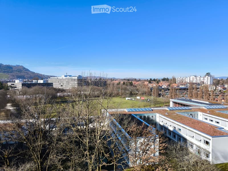 Aerial view of a building with solar panels on the roof, surrounded by trees and a grassy field, with a city skyline in the background.