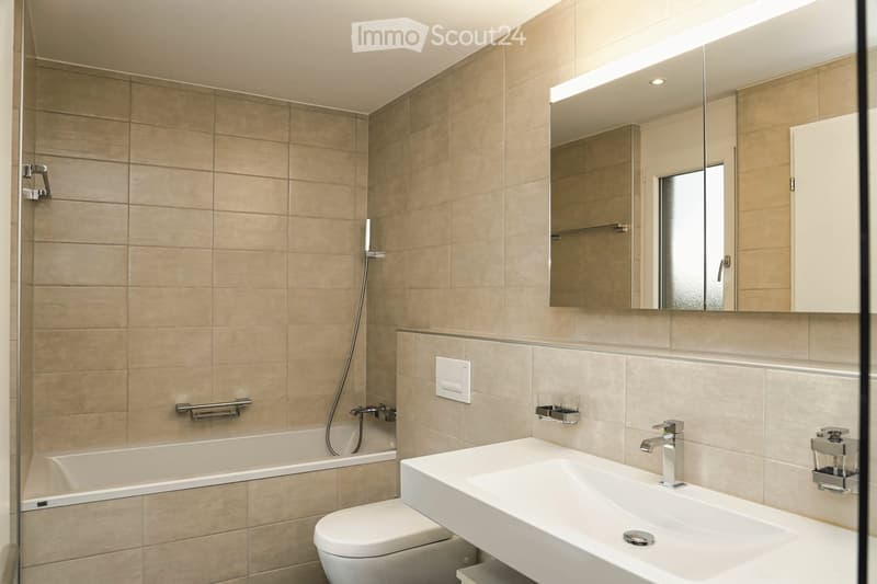Modern bathroom with beige tiled walls, a bathtub, a toilet, a large sink, a mirror with built-in lighting, and a shower hose.