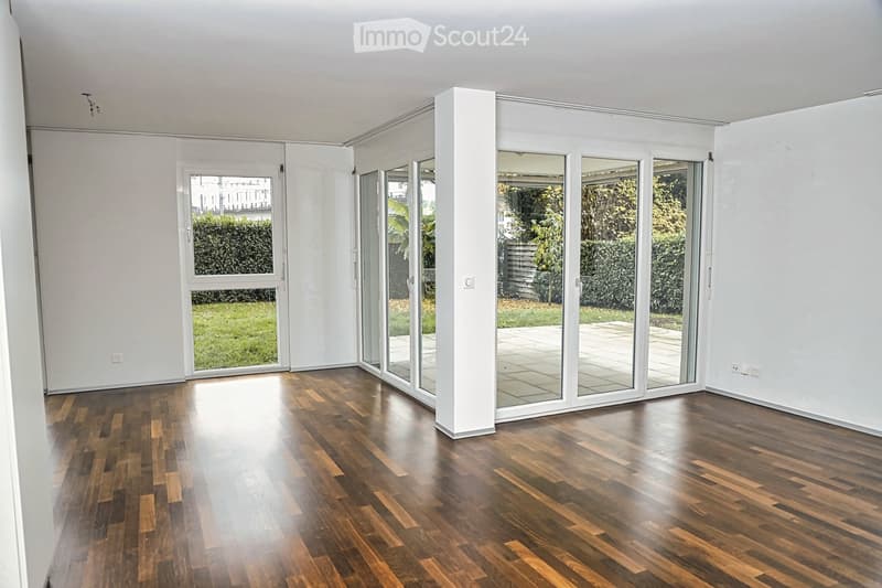 Empty room with wooden flooring, large sliding glass doors leading to a garden, white walls, and a central column.