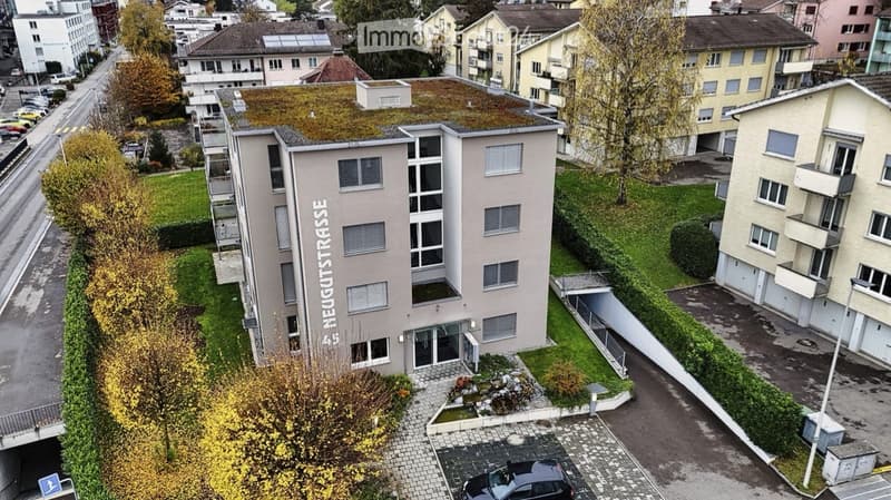 Multi-story residential building with modern facade, green roof, and surrounding greenery. Includes parking area and landscaped entrance.
