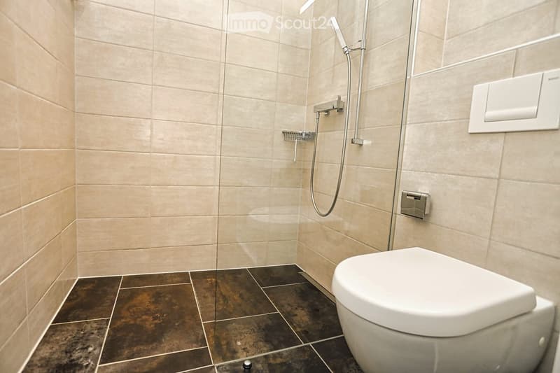 Modern bathroom with beige tiled walls, dark brown tiled floor, white toilet, and a showerhead with a rail.