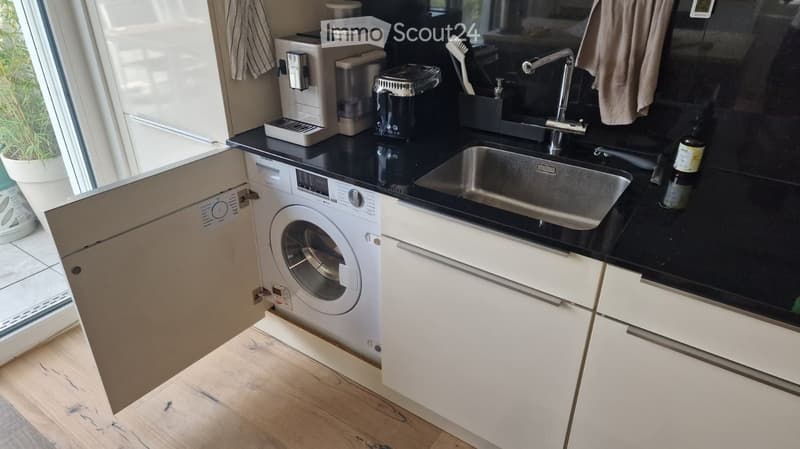 Modern kitchen with white cabinets, black countertops, stainless steel sink, and a built-in washing machine. Coffee maker and blender on the counter.