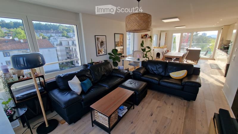 Modern living room with large windows, black leather sofa, coffee table, floor lamp, and dining area visible in the background.
