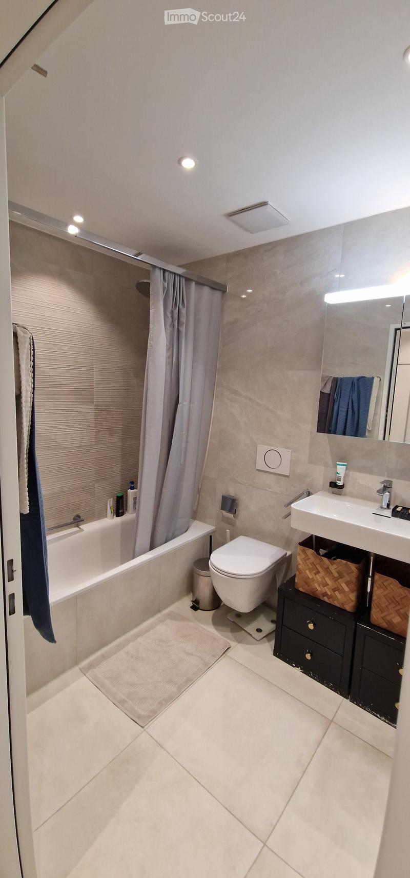 Modern bathroom with a bathtub, toilet, sink, mirror, and a black cabinet with a woven basket on top. The room features light-colored tiles and a sleek design.