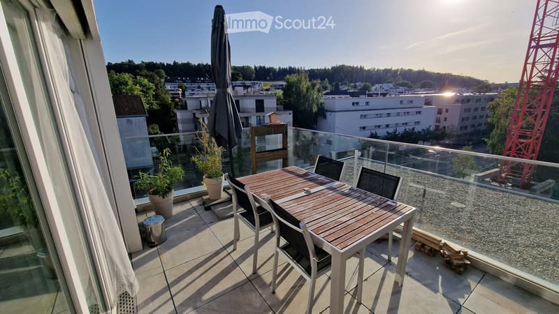 Balcony with a wooden table, chairs, umbrella, and potted plants, overlooking a cityscape with buildings and trees.