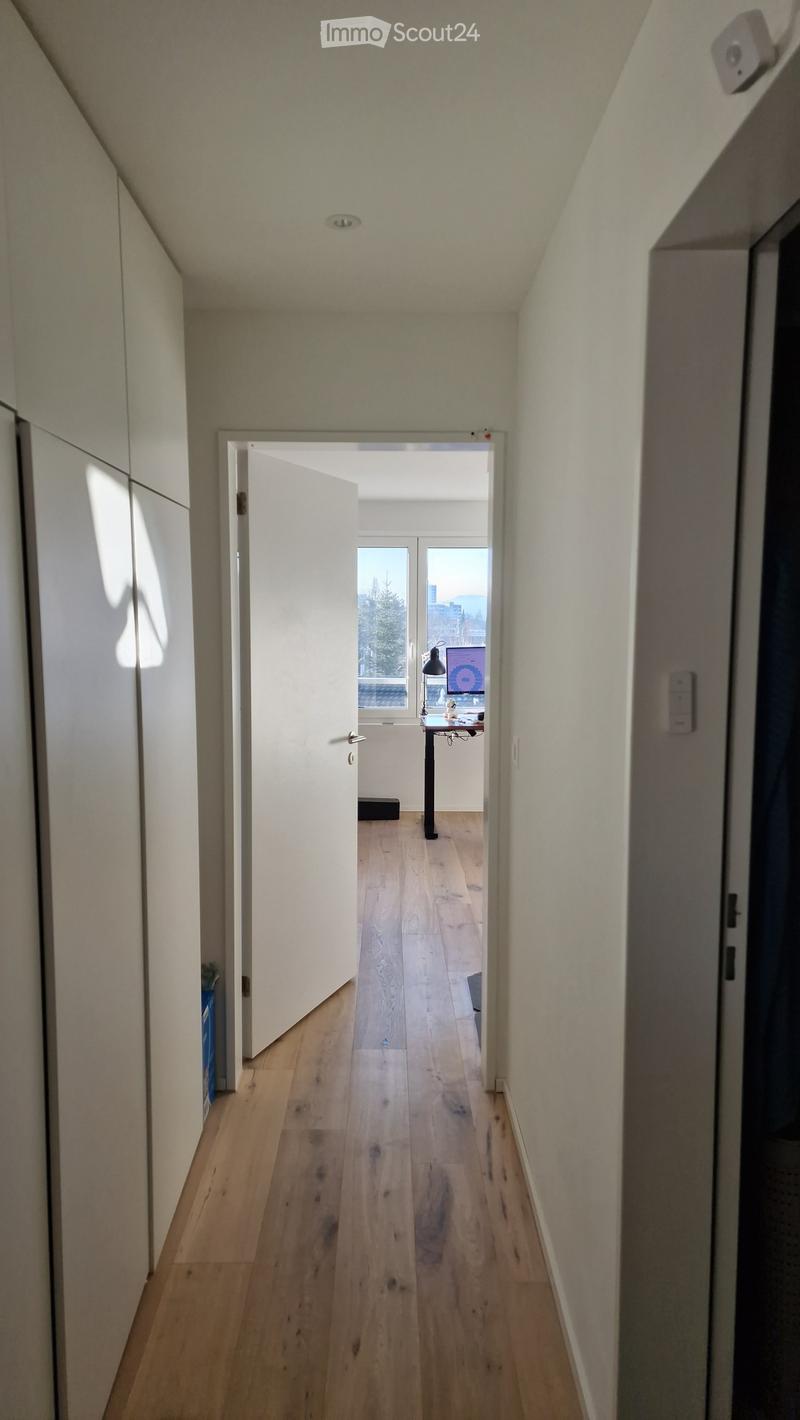 A narrow hallway with wooden flooring, white walls, and a partially open door leading to a room with a desk and a window view.