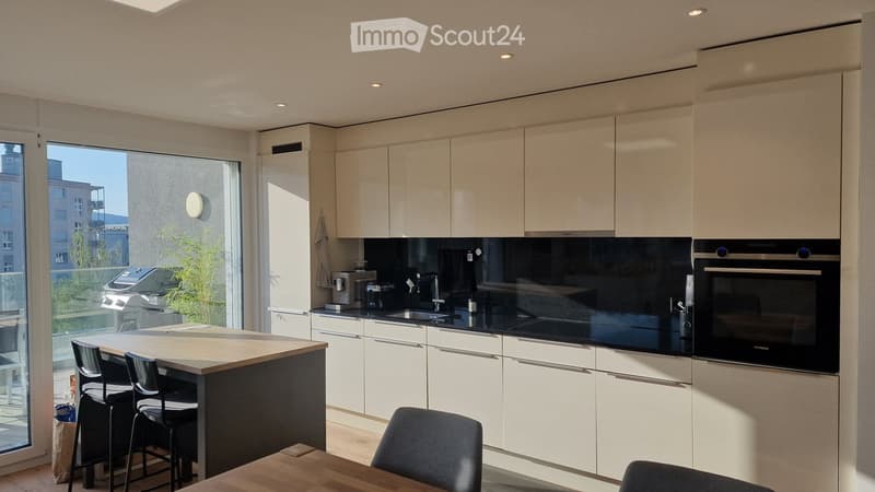Modern kitchen with white cabinets, black countertops, and a central island with bar stools. Features a built-in oven, sink, and ample natural light from large windows.