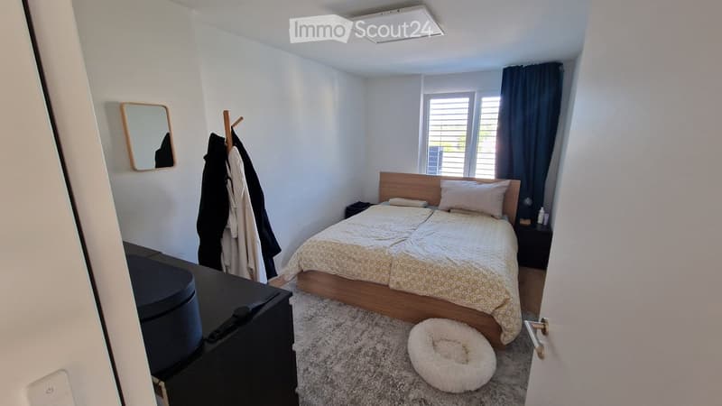 A modern bedroom with a double bed, nightstand, mirror, and a dog bed on the floor. The room features white walls, a large window with blinds, and a coat rack with jackets.