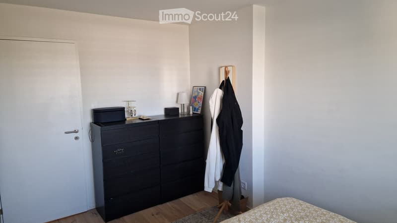 Bedroom with a wooden floor, a dark dresser with drawers, a bed with a patterned bedspread, a coat rack with jackets, a small lamp, a picture frame, and a jewelry stand.