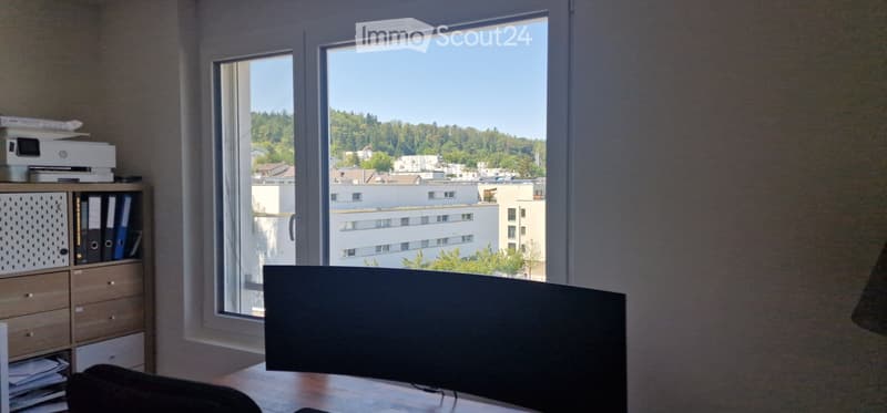 Office space with large windows overlooking a cityscape, featuring a flat-screen screen on a desk, a wooden cabinet with files and a printer, and a view of buildings and greenery.
