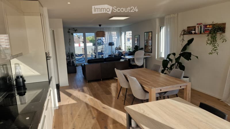 Open-plan living and dining area with wooden flooring, large windows, and a balcony. Features a sofa, dining table with chairs, and decorative plants.