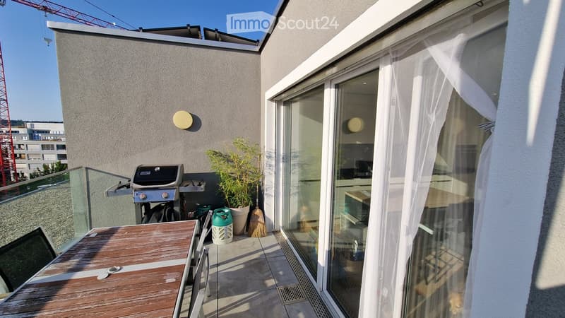 Balcony with wooden table, grill, potted plant, and sheer white curtain leading to interior space.