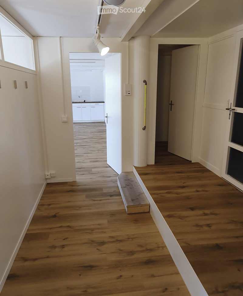 Empty hallway with wooden flooring, white walls, and a small bench placed near the wall. The hallway leads to an open doorway revealing a kitchen area with white cabinets and a countertop.
