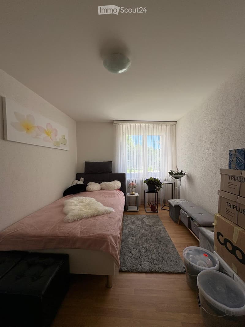 A cozy bedroom with a double bed, pink bedding, a nightstand, a small table with a lamp, a rug, and several stools and boxes stacked against the wall.