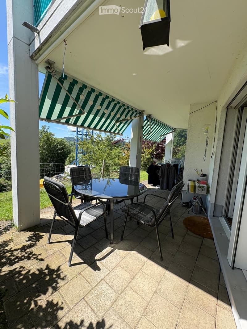 Balcony with tiled floor, glass table, four chairs, green and white striped awning, and a view of greenery.