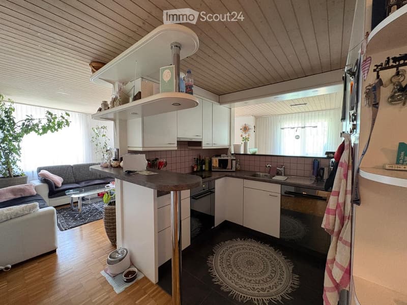 A modern, open-plan living and kitchen area with a grey sofa, coffee table, kitchen island, microwave, and various kitchen appliances. The space features wooden flooring, white cabinets, and a hanging lamp.