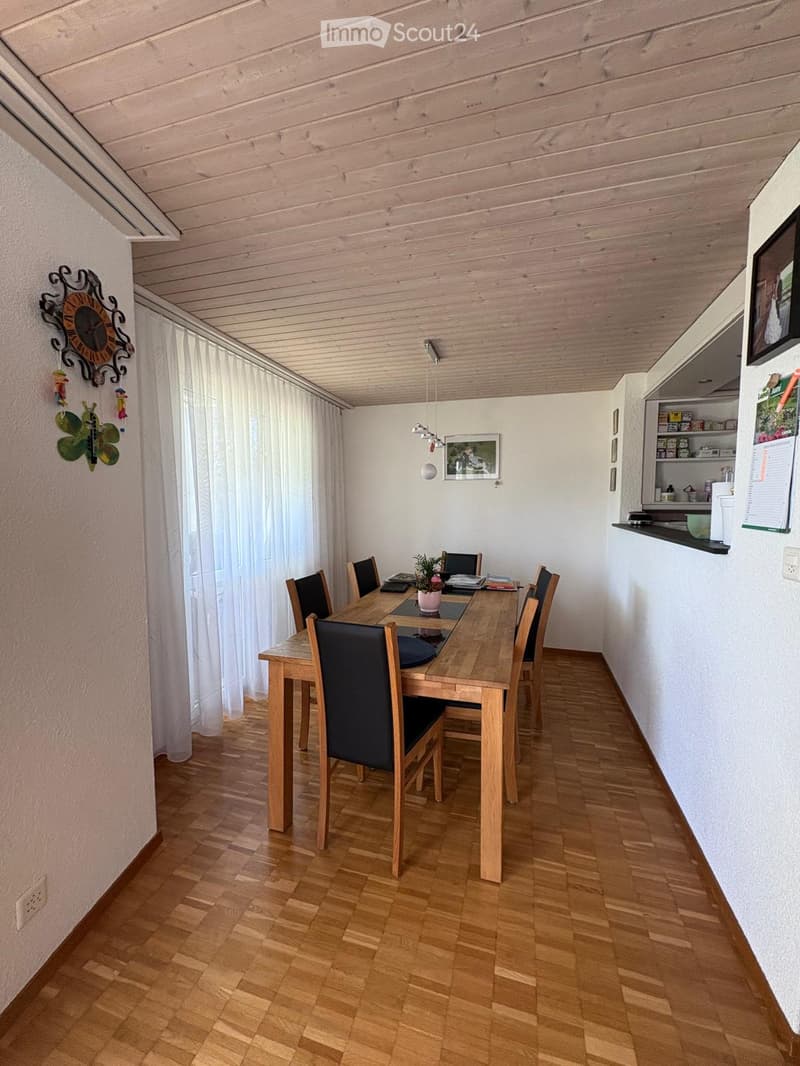 Bright kitchen with wooden flooring, a dining table surrounded by chairs, a potted plant on the table, and a small kitchen area with a fridge and cupboard.