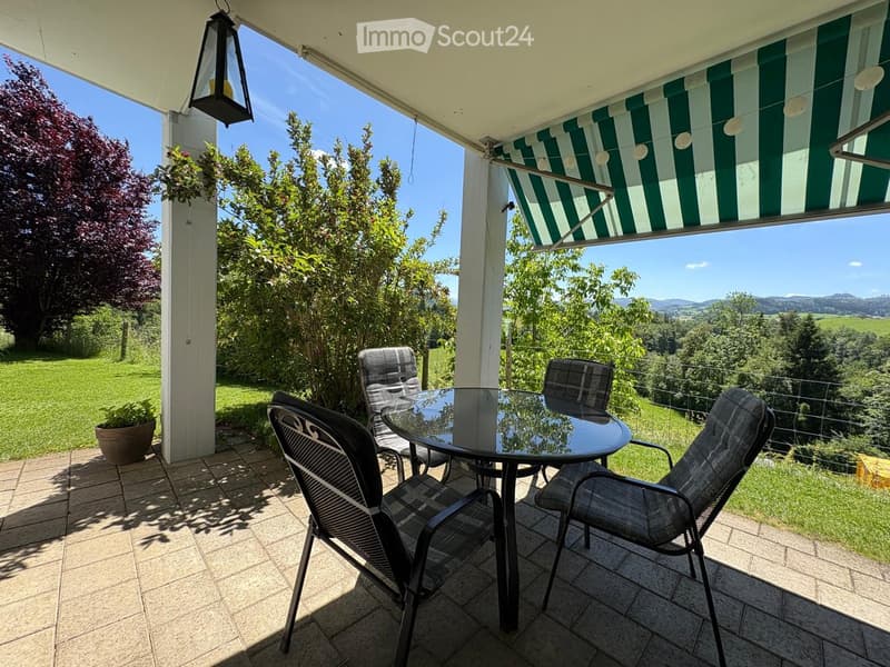 Balcony with a glass table and four chairs, overlooking a green landscape with trees and hills.