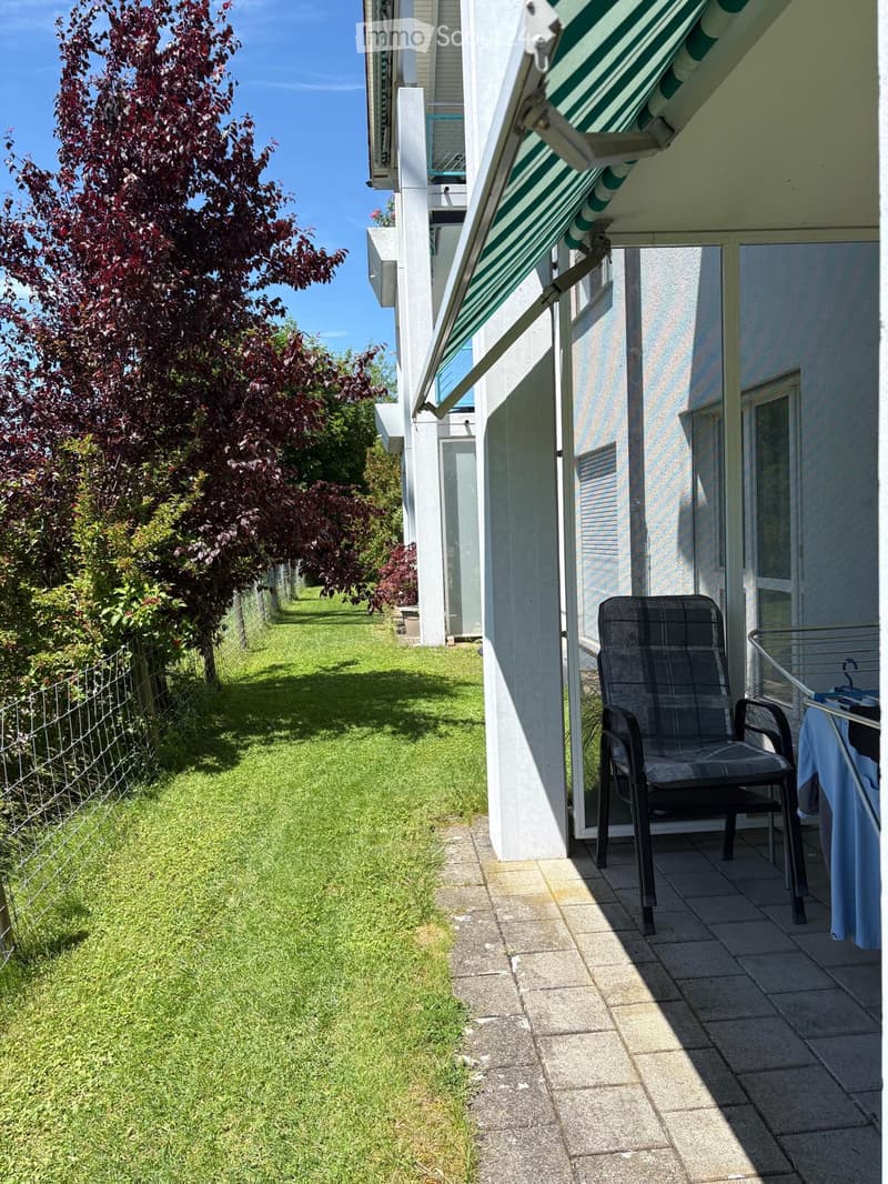 Balcony with a green and white awning, a black chair, and a small table with a blue cloth, adjacent to a well-maintained lawn and a tree.