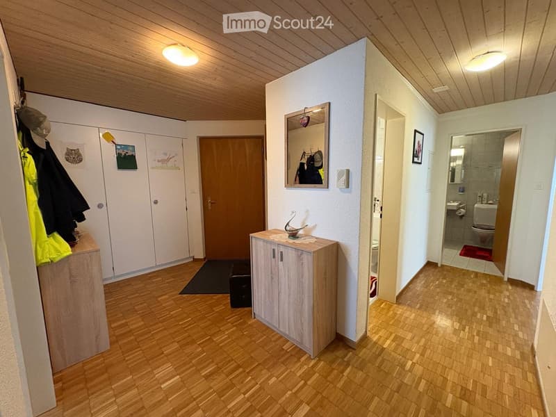 A well-lit hallway with wooden flooring, white walls, and a small wooden cabinet with a decorative item on top. The hallway leads to a bathroom with a toilet, sink, and mirror.