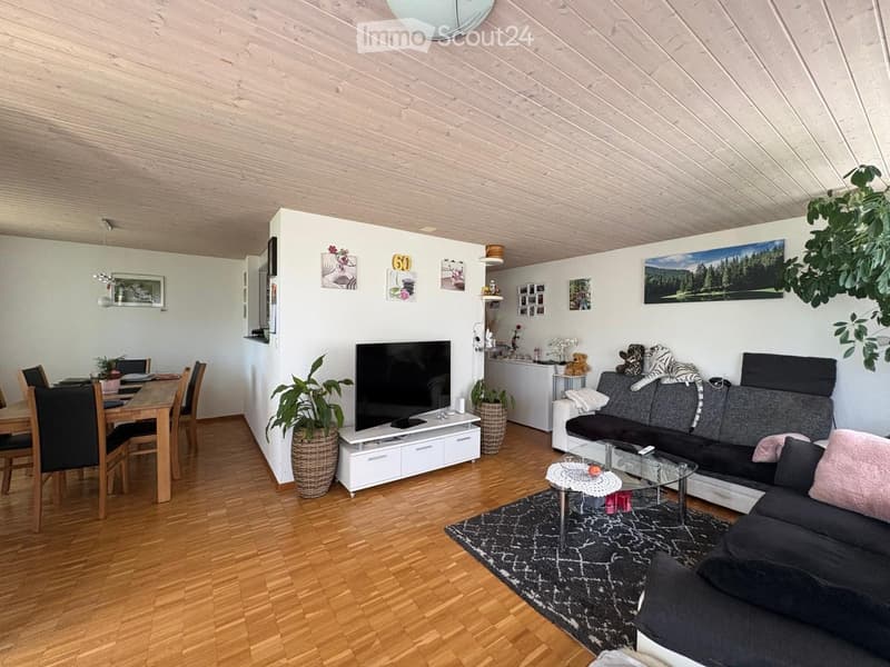 Modern living room with wooden flooring, a grey sectional sofa, a glass coffee table, a dining area with a wooden table and chairs, and a television on a white stand. The room features several potted plants and picture frames on the walls.