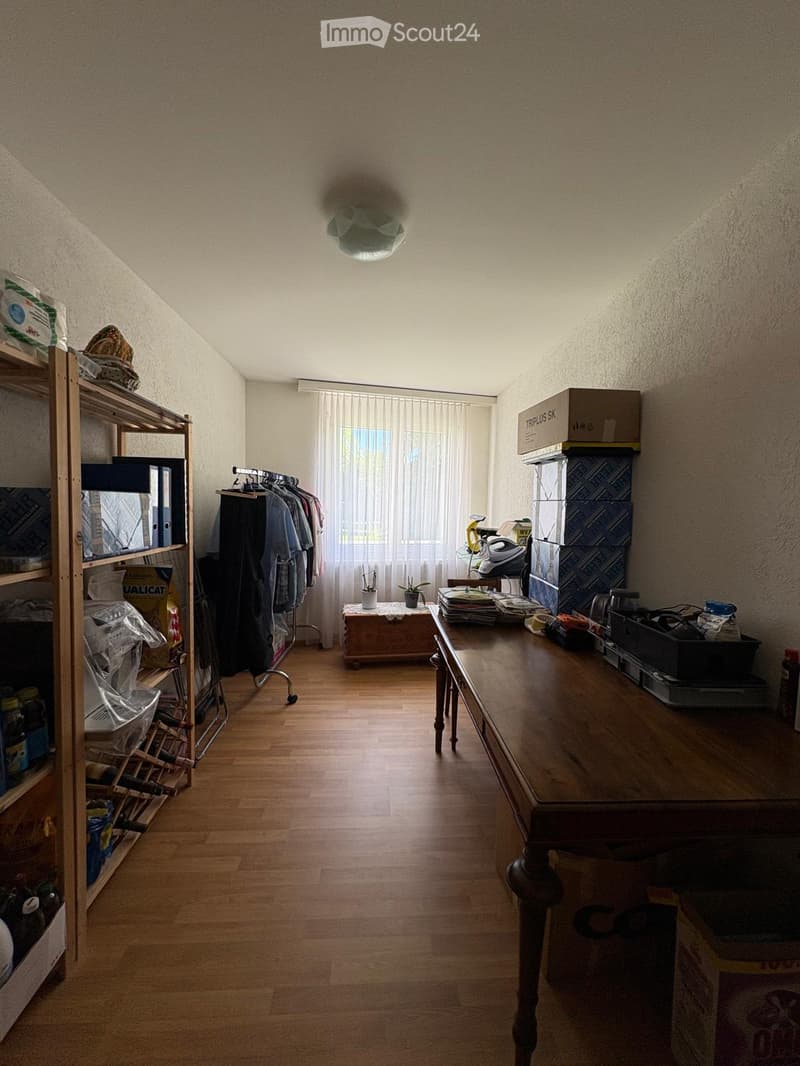 A furnished room with wooden flooring, a large window with curtains, a wooden table, a shelf, a clothing rack, and some boxes.