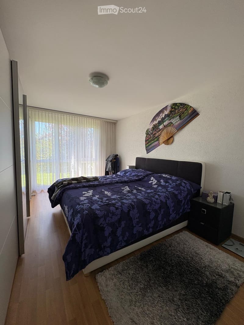 A modern bedroom with a large bed, dark headboard, nightstand, and a decorative wall fan. The room features wooden flooring, a large window with sheer curtains, and a fluffy rug.