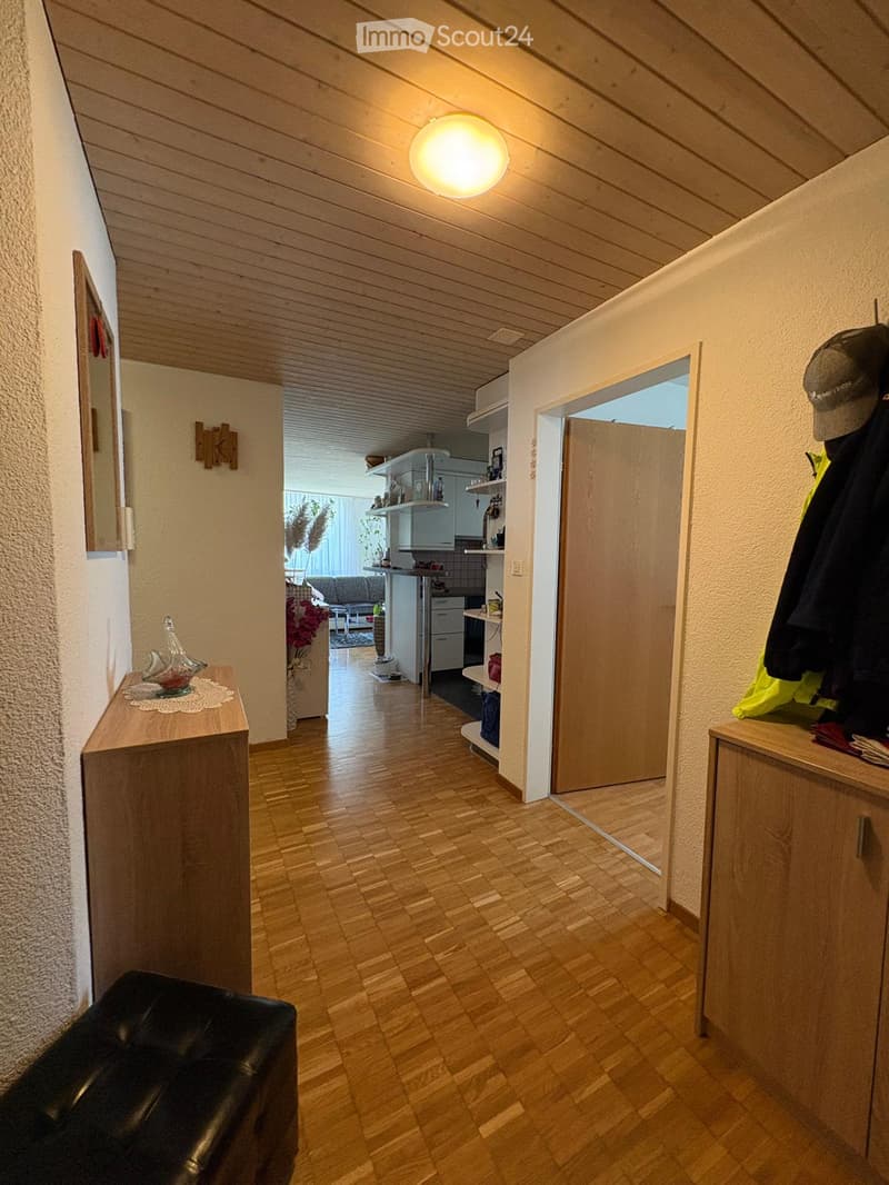 A well-lit hallway with wooden flooring, leading to a kitchen and living area. Features include a small wooden console table with a decorative glass vase, a coat rack with jackets, and a mirror on the wall.