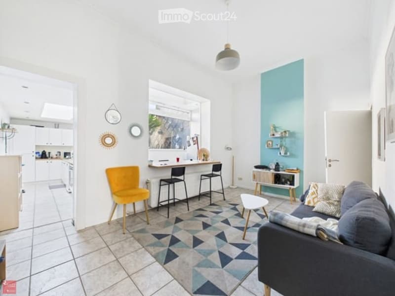 Bright, modern living space with a grey sofa, coffee table, yellow accent chair, and small kitchen area with bar stools. Features a geometric patterned rug and white tiled flooring.