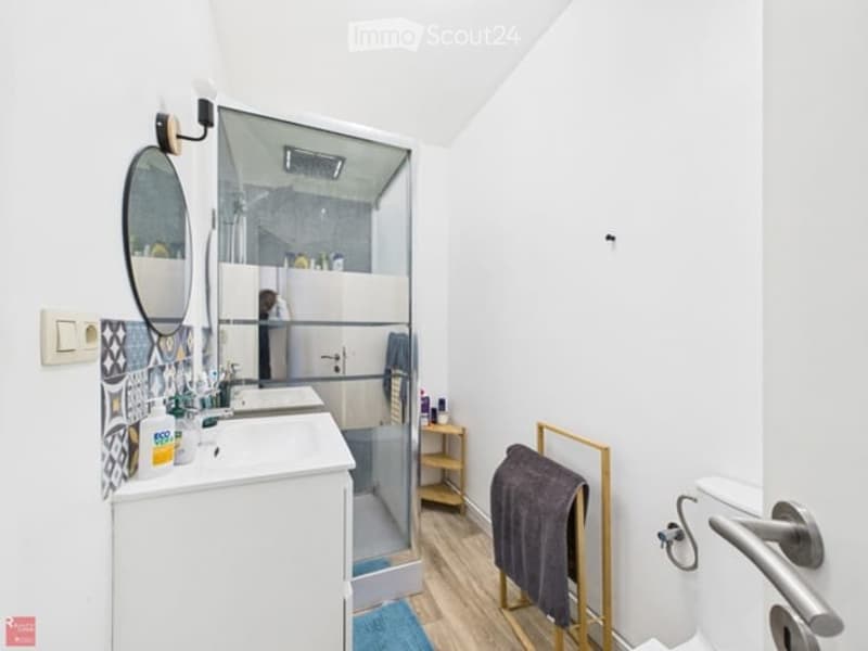 Modern bathroom with white walls, wooden flooring, a large mirror, a sink with a cabinet, a shower with a glass enclosure, a toilet, and a towel rack with a towel.
