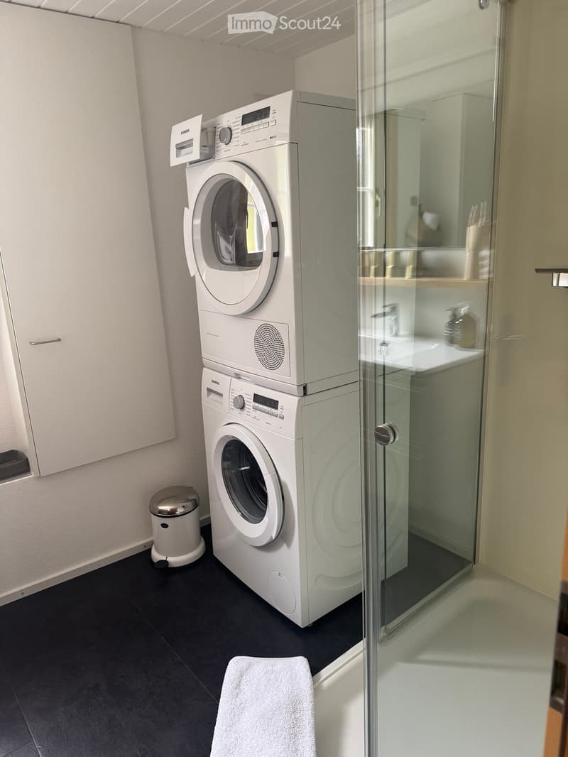 Modern bathroom with a stacked washing machine and dryer unit, a glass-enclosed shower area, a towel on the floor, and a small trash bin.