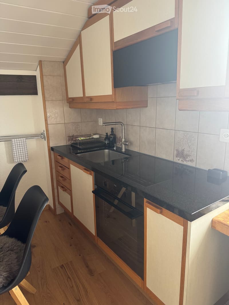 Modern kitchen with wooden cabinets, black countertop, stainless steel sink, and built-in oven. Features a sleek design with a small black chair near the counter.