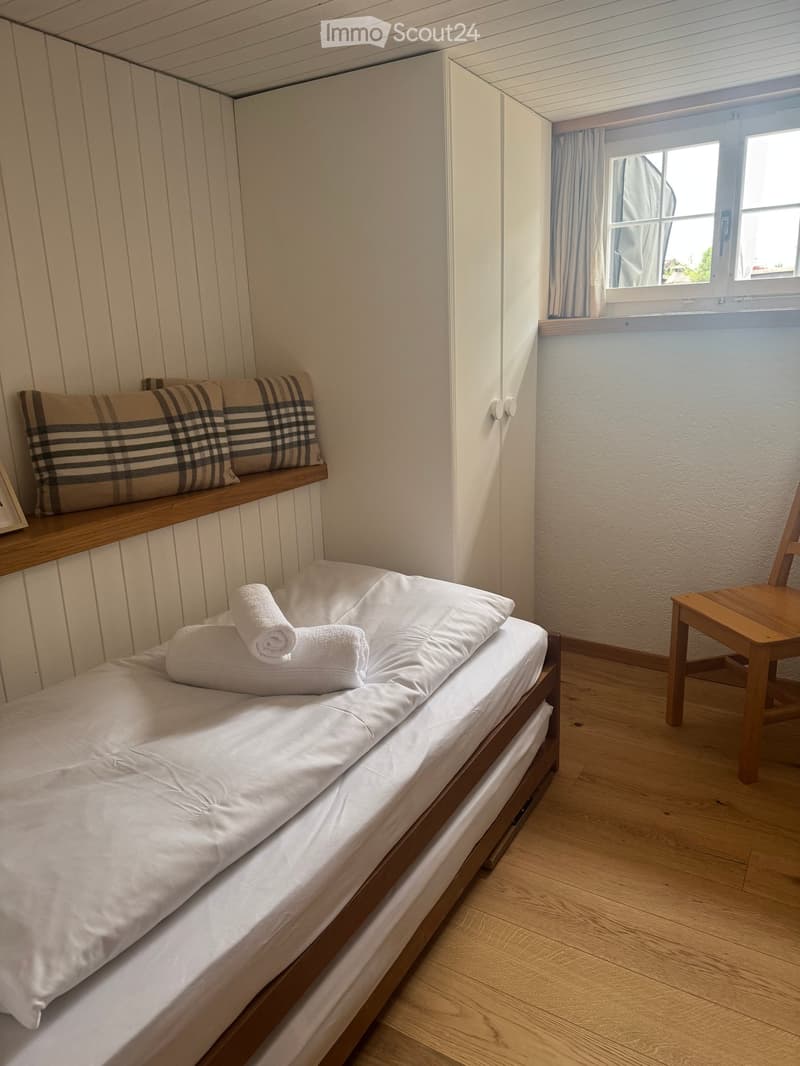 A small, neatly arranged bedroom with a single bed, pillows, towels, a wooden chair, and a shelf with additional pillows.