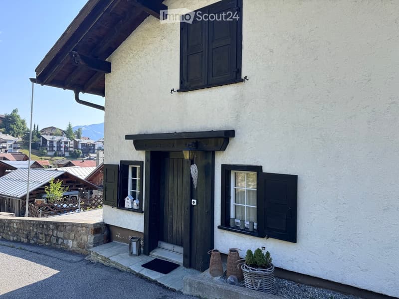 A charming two-story house with white plaster walls, dark wooden shutters, and a wooden entrance door. The entrance features a small porch with a lantern and decorative wicker baskets. The surrounding area includes a stone wall, a small garden with potted