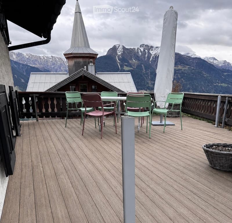 Wooden deck with a table and chairs, overlooking a mountain landscape with a church spire visible in the distance.