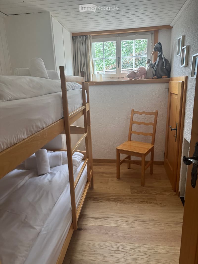 Cozy bedroom with a wooden bunk bed, a small wooden chair, and decorative toys on the windowsill. Light wooden flooring and white walls.